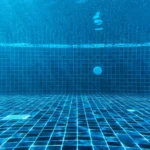 An underwater view of a blue tiled swimming pool with a drain